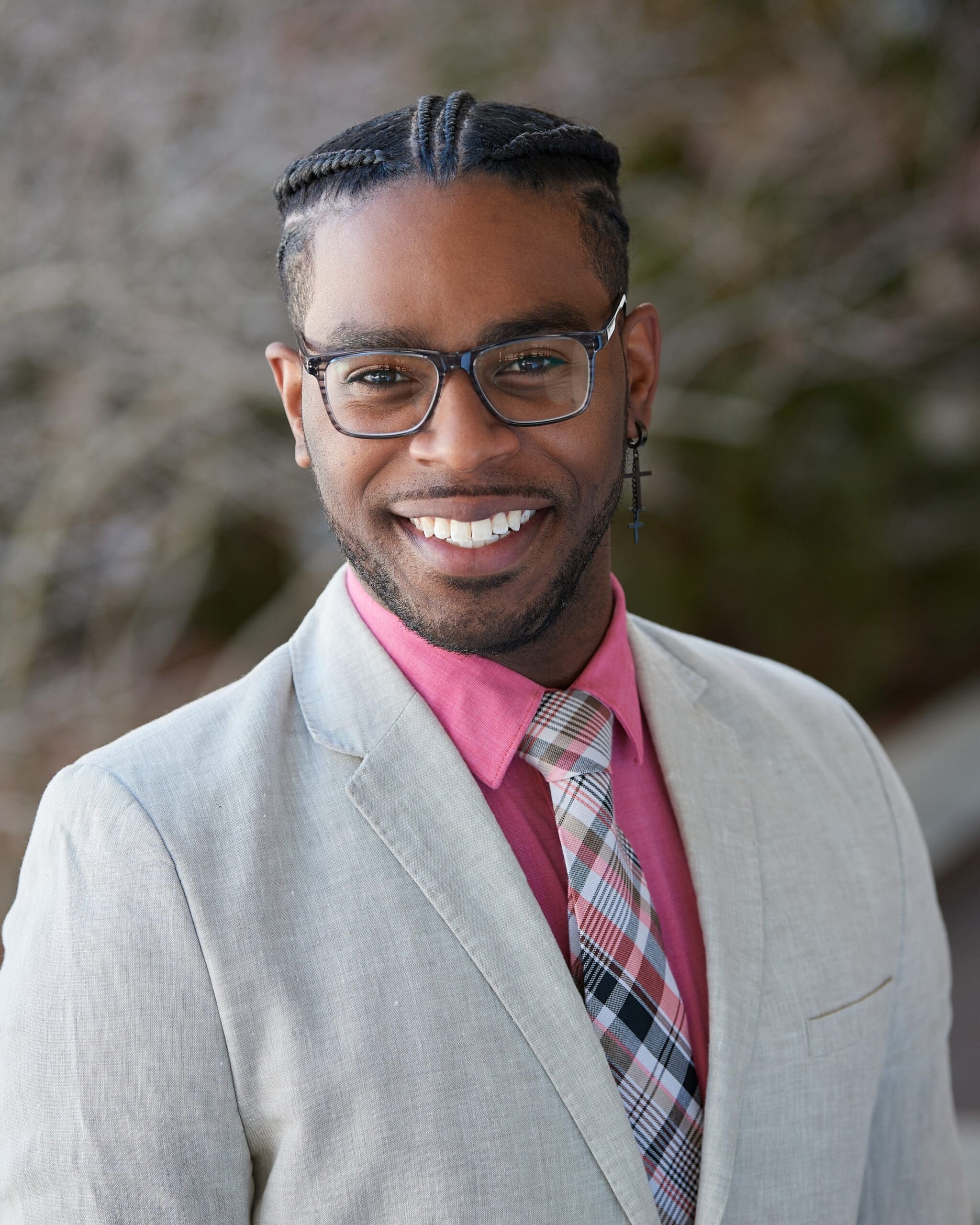 Smiling man in a light gray blazer, pink dress shirt, plaid tie, wearing glasses outdoors.