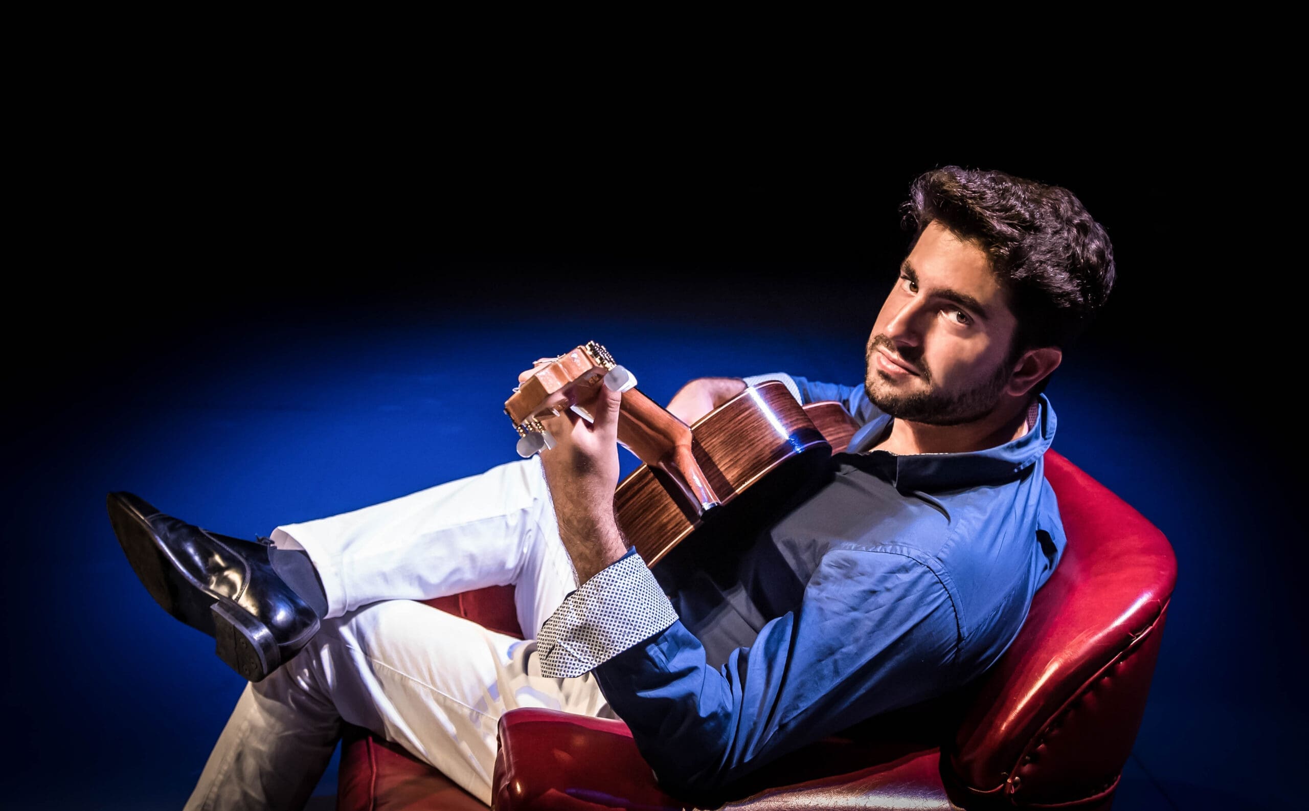 Man wearing a blue shirt sits in a red chair, playing an acoustic guitar on a dark stage.Stage lighting casts blue hues on the performer and guitar.
