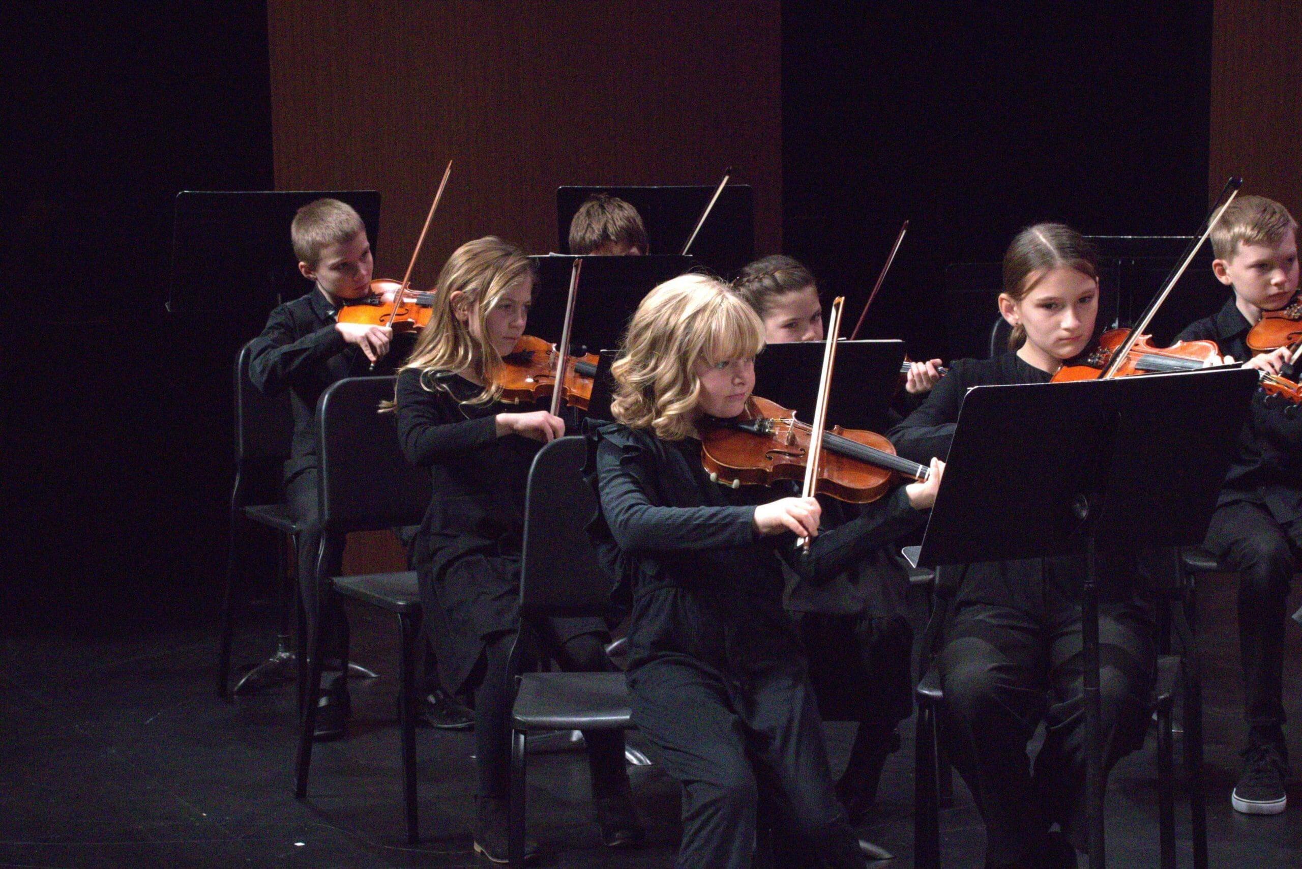 Young students in black perform a violin concert on stage, focused on their instruments in a youth orchestra setting.