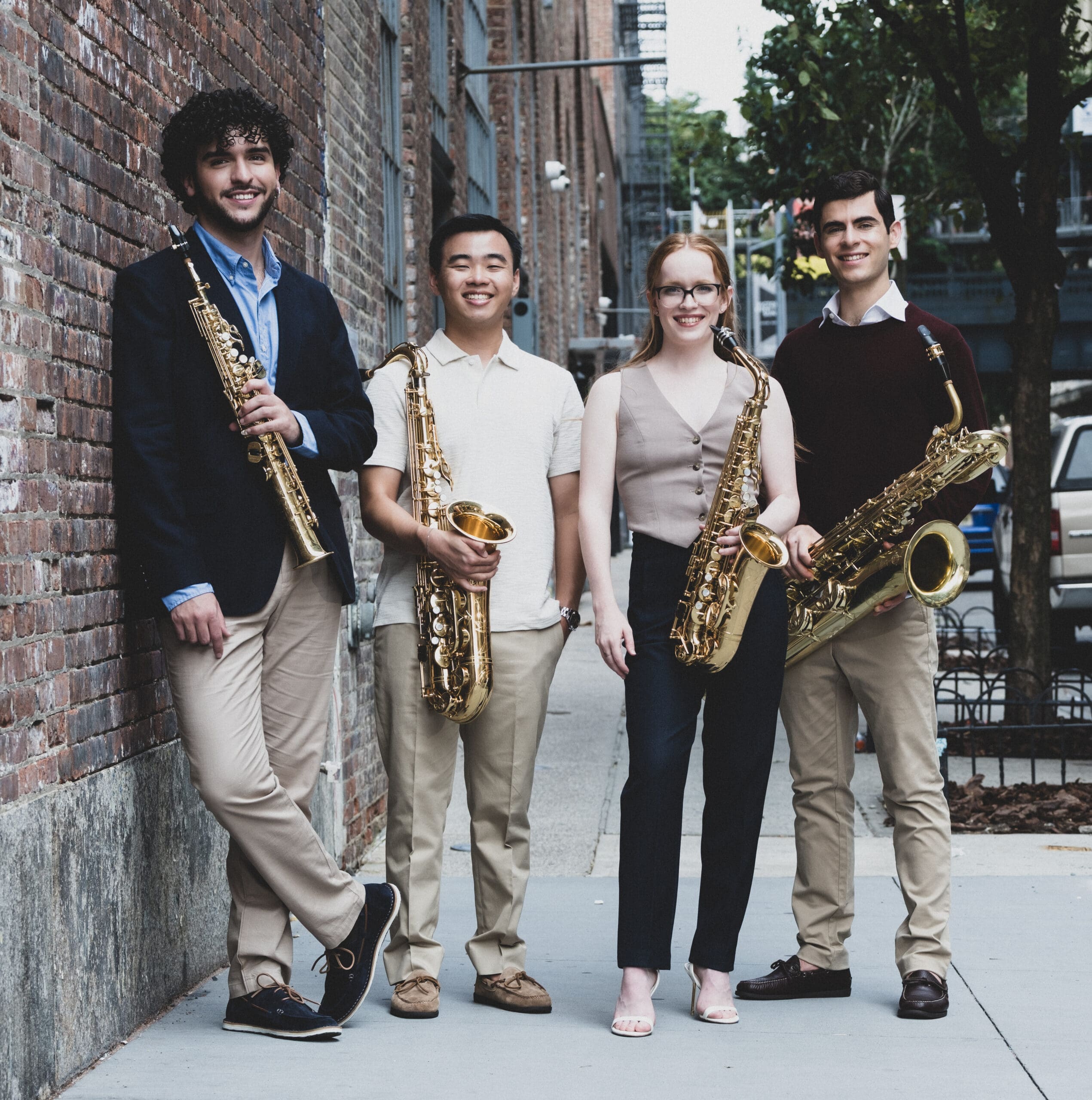 Four young adults stand on a city sidewalk against a brick wall, each holding a saxophone and smiling at the camera.