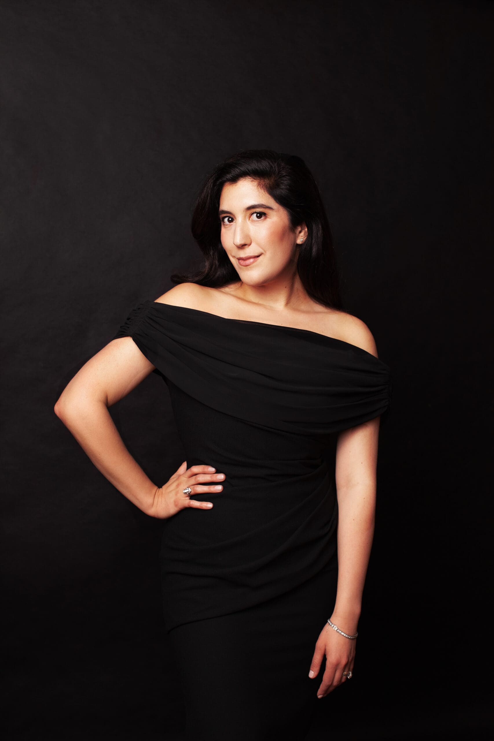 Portrait of a woman in an off-the-shoulder black dress standing against a dark studio background, hand on hip and looking at the camera.
