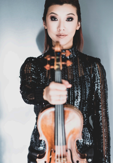 Female violinist in a sparkly black outfit extends a violin toward the camera, portrait shot with a soft background.